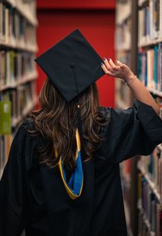 a woman wearing a graduation cap and gown walks through a library with bookshelves