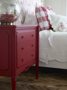 a red chest of drawers sitting in front of a bed with white sheets and pillows