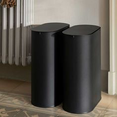 two black trash cans sitting next to each other in front of a radiator