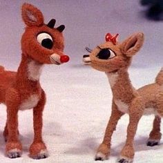 two stuffed deers standing next to each other on a snow covered ground with trees in the background