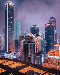 a man standing on top of a roof in front of tall buildings at night time