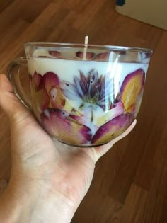 a hand holding a glass cup with flowers on it and a candle in the middle