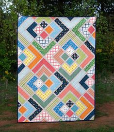 a multicolored quilt is hanging on the side of a tree trunk in front of some trees