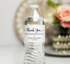 a bottle of water with a thank you label on the front and side, sitting next to a vase filled with flowers