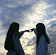 two girls are standing in front of the sky making a heart shape with their hands