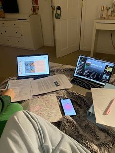 two laptops and papers on a bed with someone's feet in the air