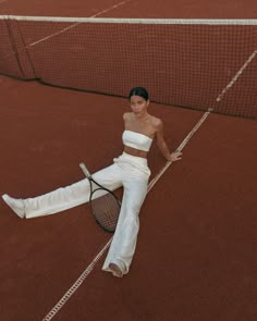 a woman sitting on the ground with a tennis racket in her hand and wearing white pants