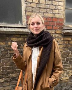 a woman standing in front of a brick building holding onto a brown purse and wearing a scarf around her neck