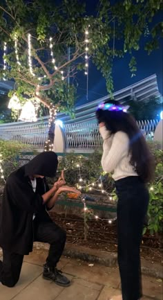 a man kneeling down next to a woman with a hood on and lights hanging from the trees