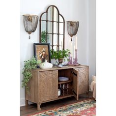 a wooden cabinet with plants and candles on it in front of an ornate mirror above