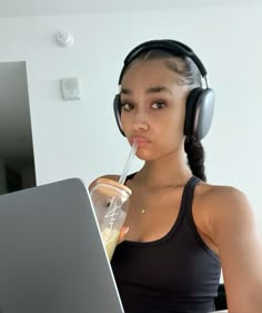 a young woman wearing headphones drinking from a cup while using a laptop computer on her desk