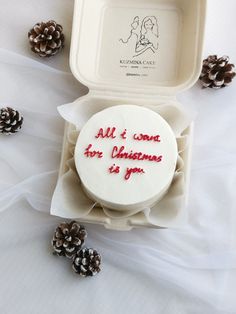 a white cake in a box with red writing on it and pine cones around it