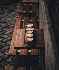 three cups of coffee sit on a wooden table with benches in the background and cobblestone pavement