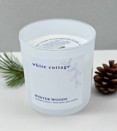 a white candle sitting next to a pine cone on top of a table with the words winter woods printed on it