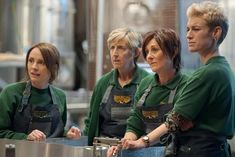 three women in green aprons are standing behind a counter and looking at the camera