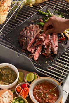 someone is grilling steak on the grill with vegetables and dipping sauces around it