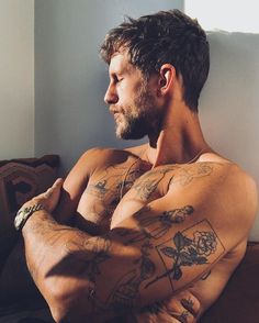 a man with tattoos on his arms and chest sitting in front of a window looking off into the distance