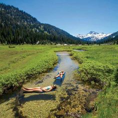 two people laying on their stomachs in the middle of a stream with mountains in the background