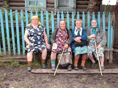 three older women sitting on a bench in front of a blue fence and wooden house