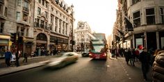 a double decker bus driving down a street next to tall buildings with people walking on the sidewalk