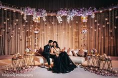 a man and woman sitting on a couch in front of a stage with flowers hanging from the ceiling