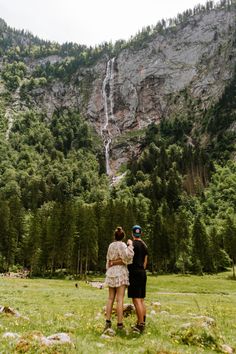 two people standing in front of a waterfall with trees on the side and mountains behind them