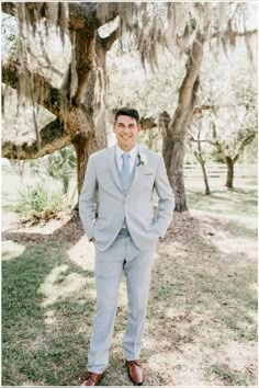 a man standing in front of a tree wearing a gray suit and blue tie with his hands in his pockets