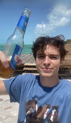 a young man is holding up a bottle of beer near the ocean and boardwalks