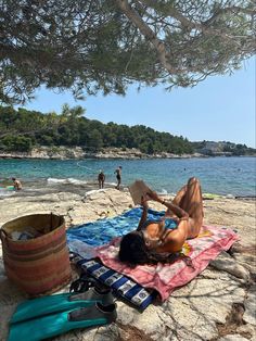 a man laying on top of a towel next to a tree near the ocean and people swimming in the water