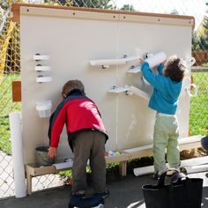 two young children are playing with cups on a white box in front of a chain link fence