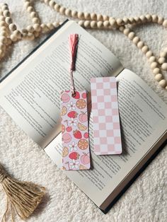 two bookmarks sitting on top of an open book next to beads and tassels