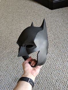 a hand holding a batman mask on top of a carpeted floor in front of a bed