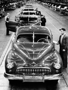 an old black and white photo of people looking at cars in a car assembly line