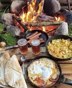 an open fire pit with food and drinks on the table next to it in front of some logs