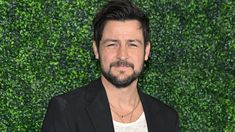 a man with a beard wearing a black suit and white shirt standing in front of a green wall