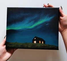 a person holding up a painting with a house in the background and aurora lights above