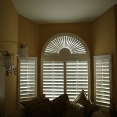 an arched window with white shutters and pillows on the couch in front of it