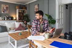 a man sitting at a table with a laptop and papers in front of him, while another person sits on the couch behind him