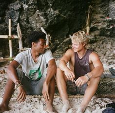 two young men sitting on the sand next to each other