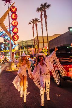 two women in white outfits and cowboy hats are walking down the street with their arms outstretched