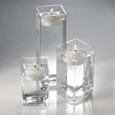 three clear glass vases with candles in them on a white surface, one is filled with water and the other has a lit candle