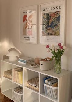 a white shelf with books and flowers on it in front of two pictures hanging on the wall