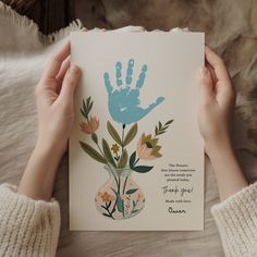 a person holding up a card with a handprint on it and flowers in a vase