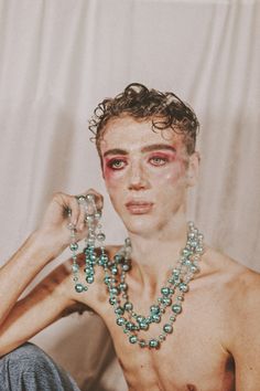 a shirtless man with beads around his neck posing for a photo in front of a white backdrop