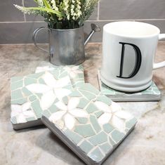 two coasters with the letter d on them next to a coffee mug and flower arrangement