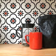 an orange coffee mug sitting next to a black bag on a wooden table in front of a patterned wallpaper