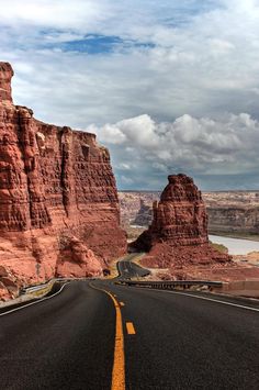 an empty road in the middle of some red rocks and water with clouds above it