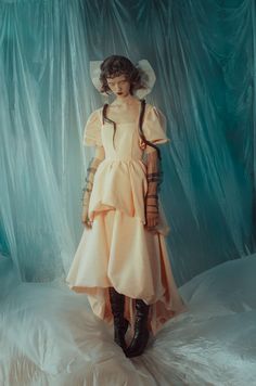 a woman in a dress and hat is standing on a bed with blue curtains behind her