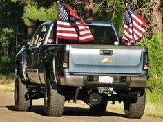 a silver truck with american flags on the back of it's bed in front of some trees