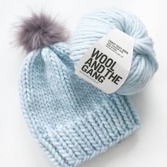 a blue knitted hat with a gray pom - pom on it and a wool and the gang tag